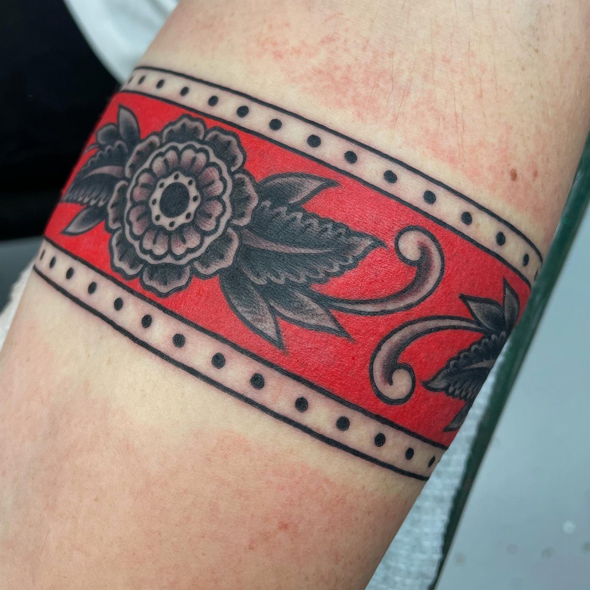 A red arm band tattoo with black floral patterns and dotted borders on a person's skin.