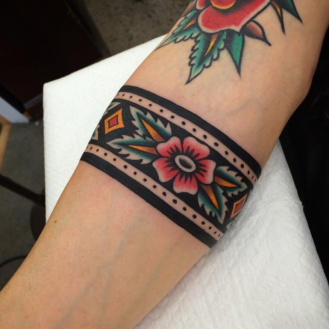 An American traditional arm band tattoo featuring a central pink flower with teal accents on a forearm.