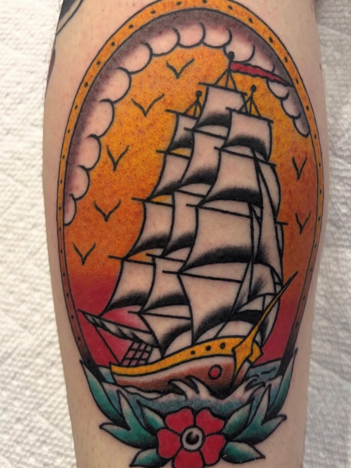 A traditional style tattoo of a sailing ship on a sunset background with waves and a rose.