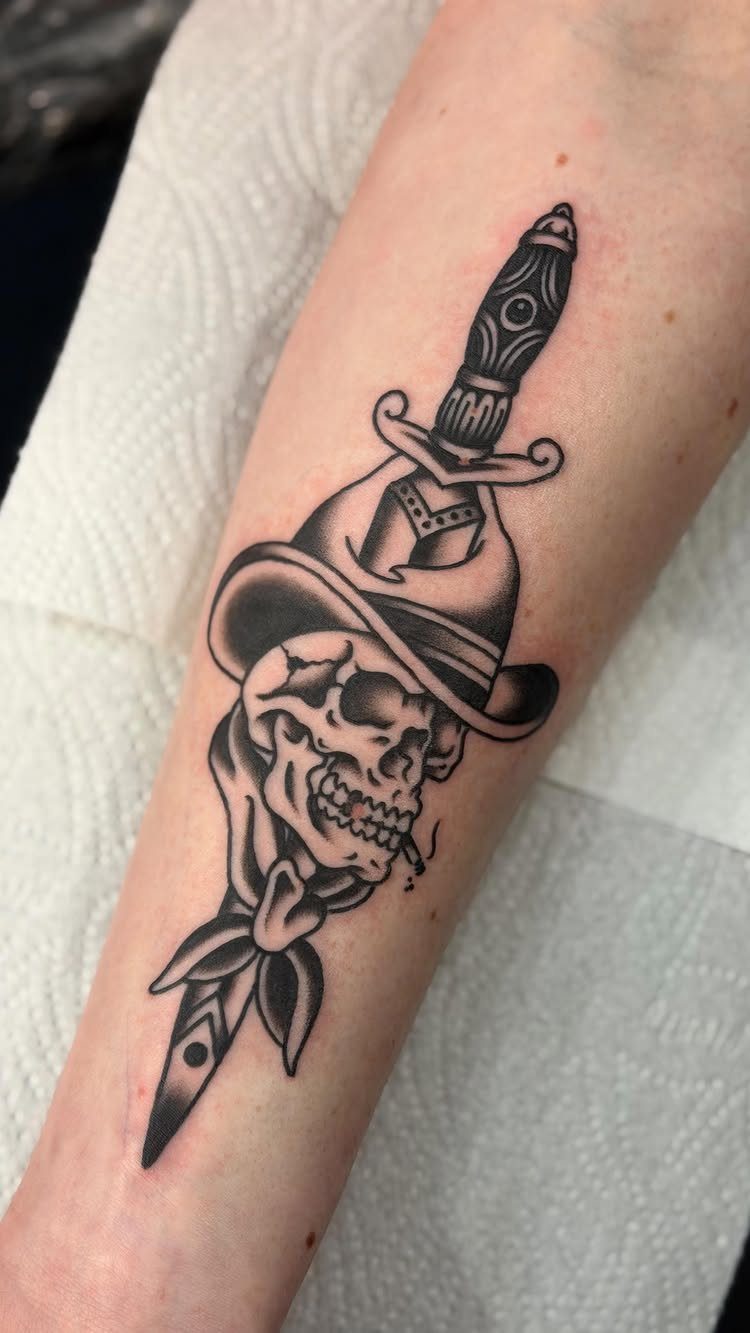 A black and grey tattoo of a skull wearing a hat with a dagger through it.