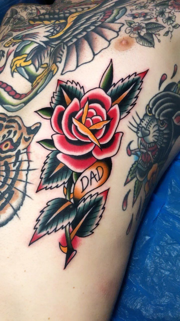 A red traditional American style rose tattoo with green leaves and a small banner saying DAD.