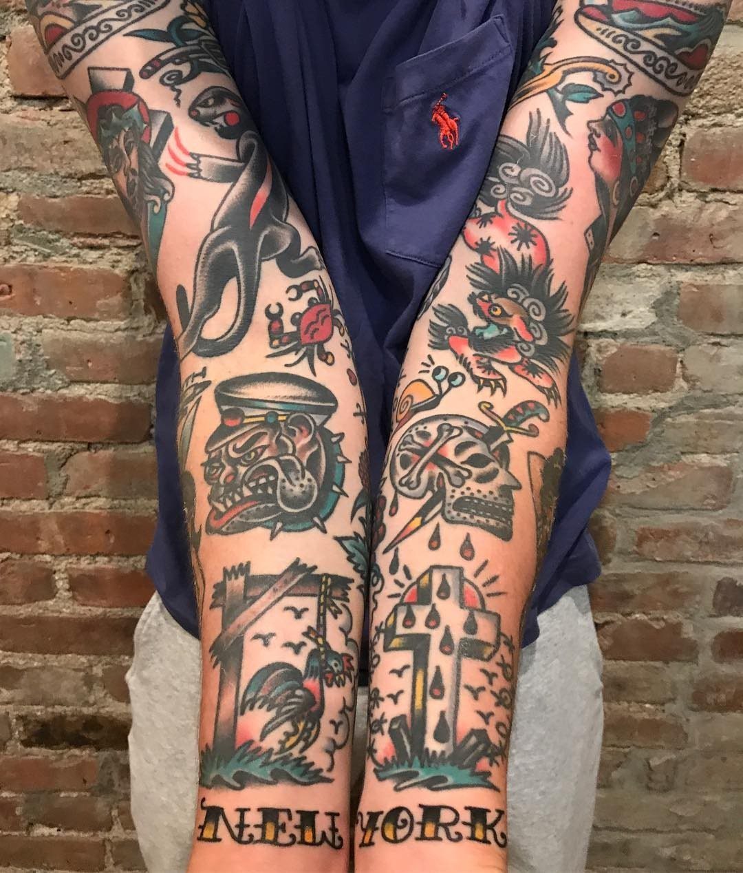 A person's arms displaying numerous colorful American traditional style tattoos against a brick wall background.
