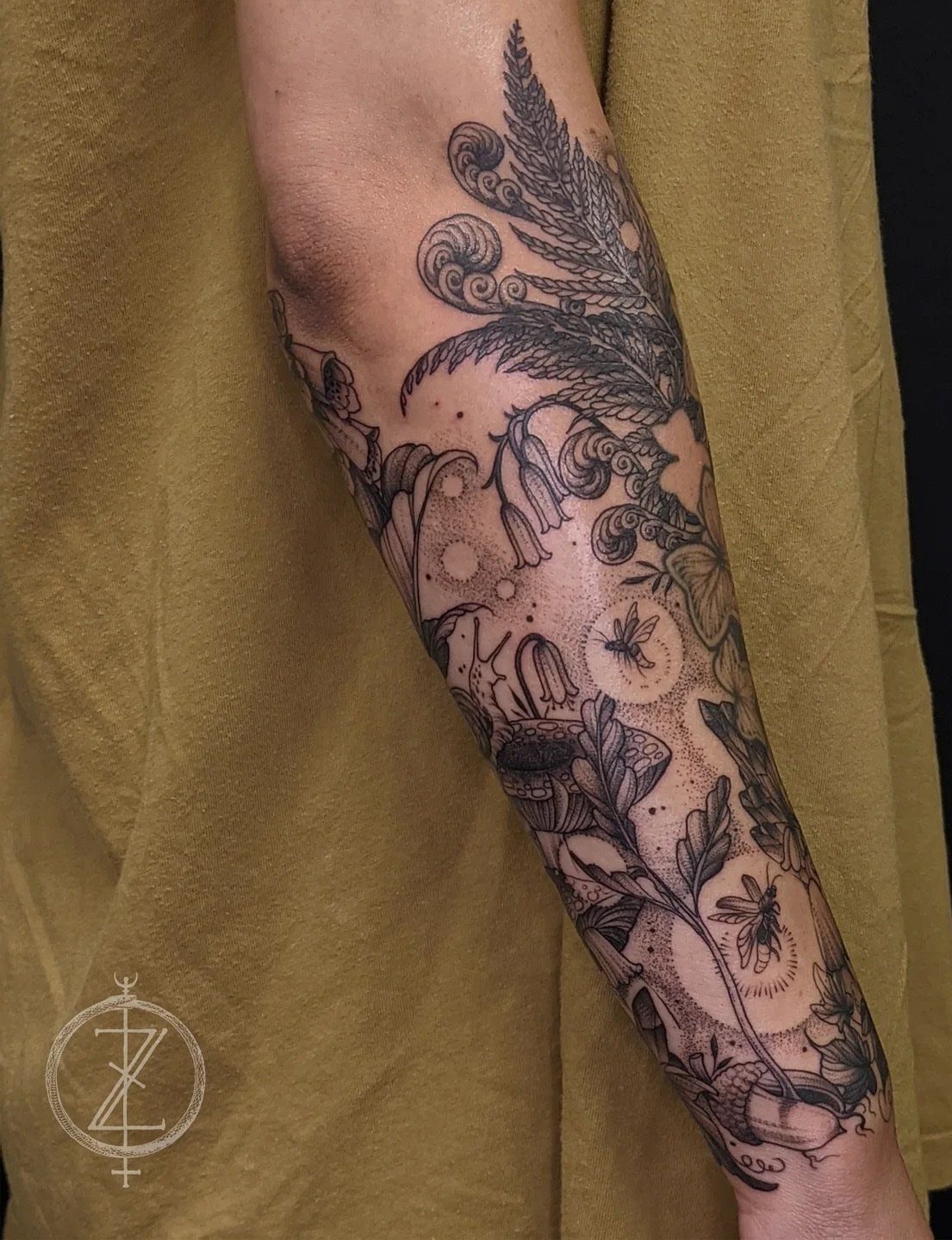 8 Arm Tattoo Filler Ideas for Sleeves That Actually Work 2 Detailed black and grey botanical tattoo featuring ferns, flowers, and bees on a person's forearm.