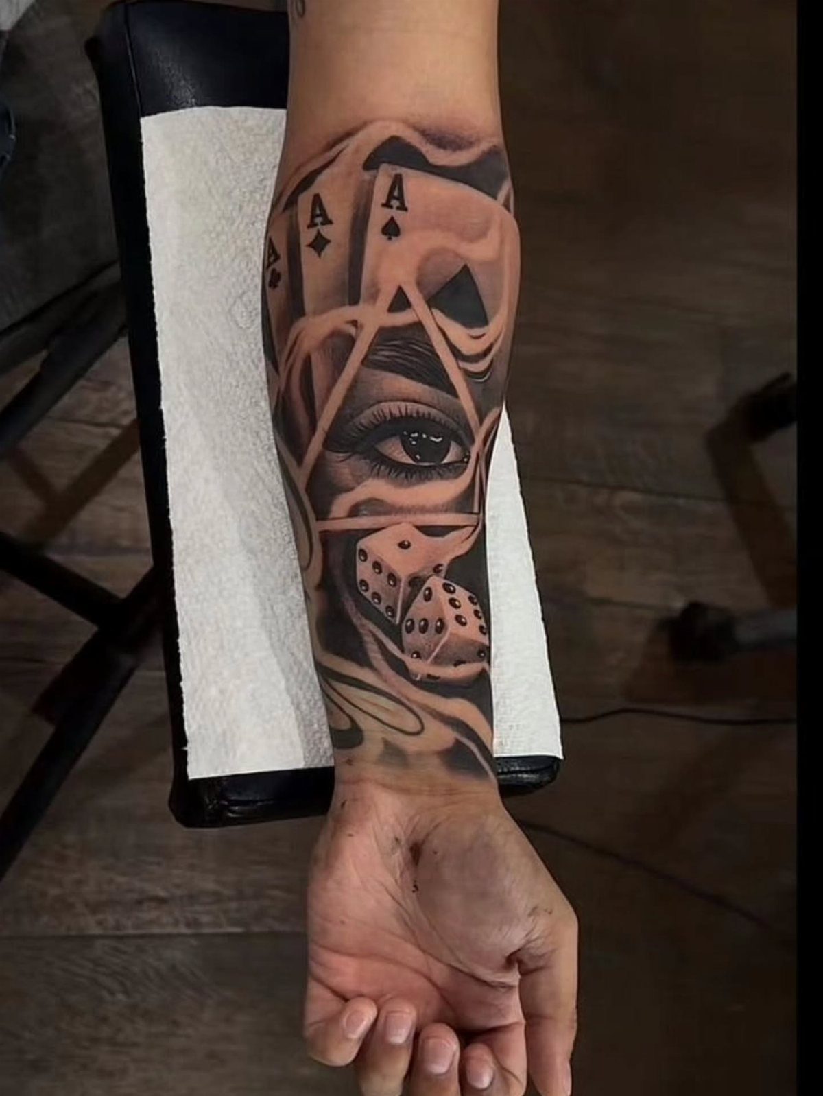 8 Arm Tattoo Filler Ideas for Sleeves That Actually Work 8 A detailed black and grey forearm tattoo depicting an eye, playing cards, and dice amidst swirling smoke patterns.