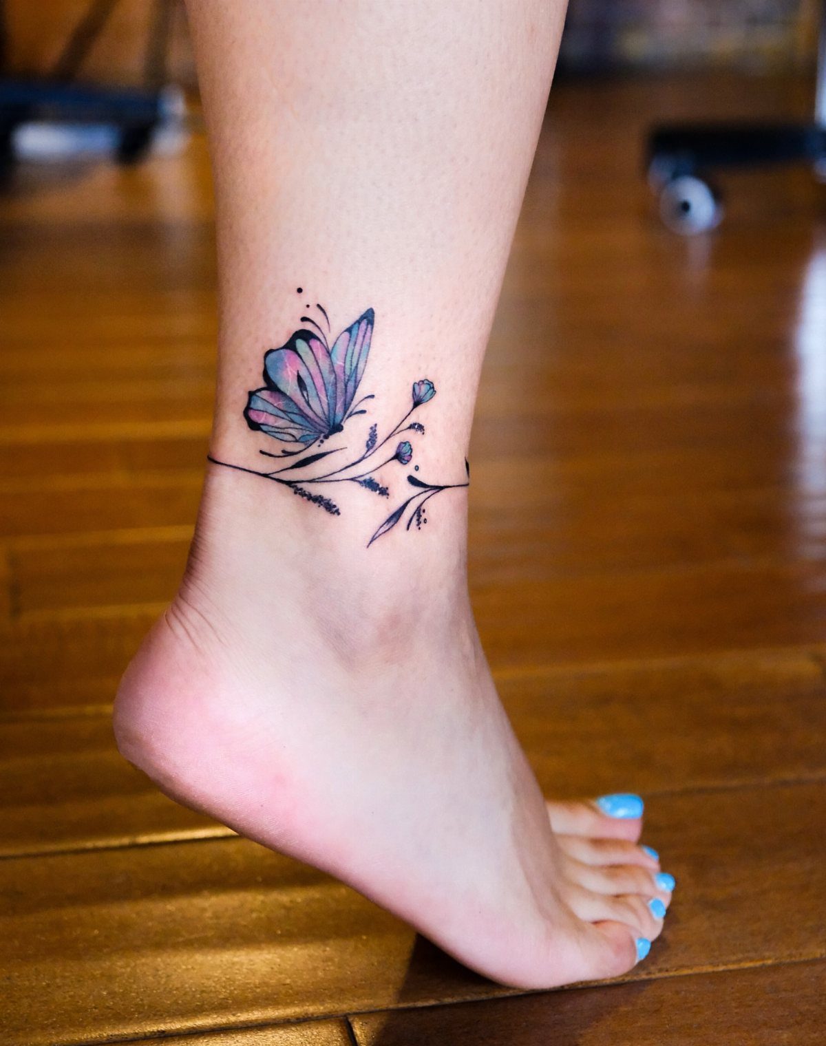 10 Around Ankle Tattoo Ideas That Follow the Body's Line 5 A butterfly tattoo with blue wings and floral accents wrapped around a person's ankle.