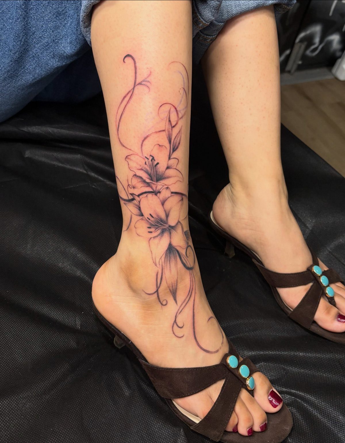 10 Around Ankle Tattoo Ideas That Follow the Body's Line 1 A large black and grey floral tattoo with lilies and vines on a person's lower leg.