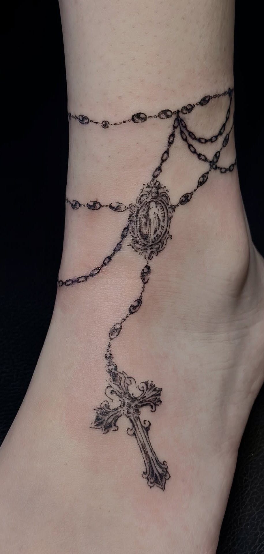 10 Around Ankle Tattoo Ideas That Follow the Body's Line 4 A black ink rosary tattoo encircling a person's ankle with a hanging cross.