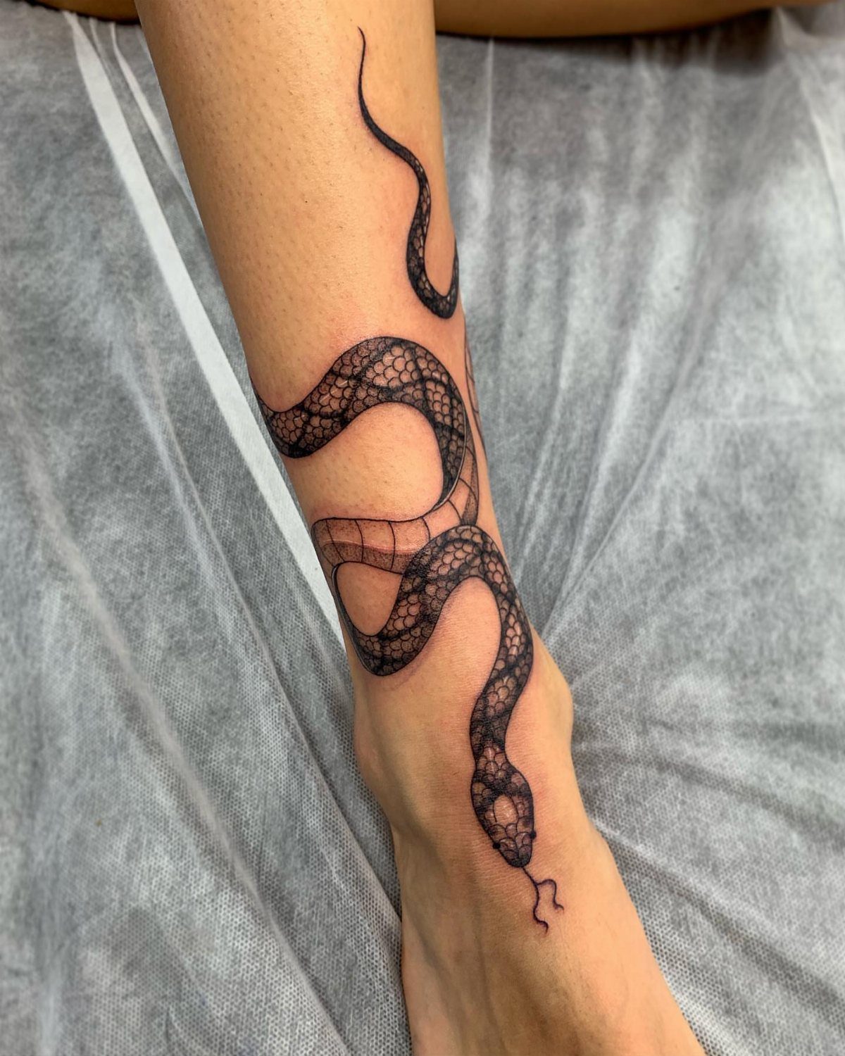 10 Around Ankle Tattoo Ideas That Follow the Body's Line 3 A black ink snake tattoo winding around a person's lower leg and ankle.