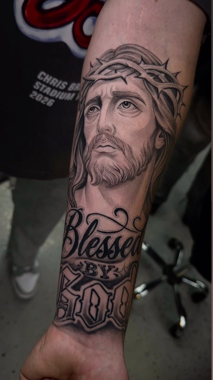 A black and grey realism tattoo of Jesus Christ with text on a forearm.