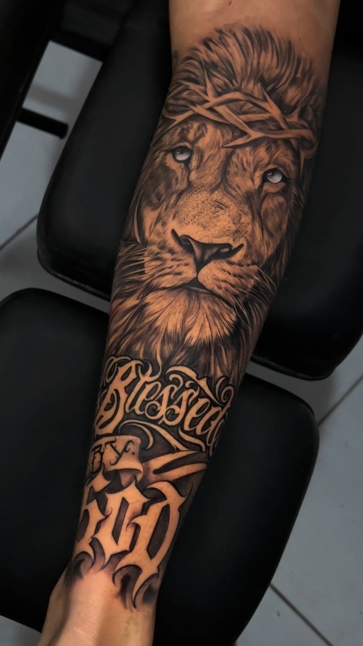 A detailed black and grey forearm tattoo of a lion wearing a crown of thorns with script.