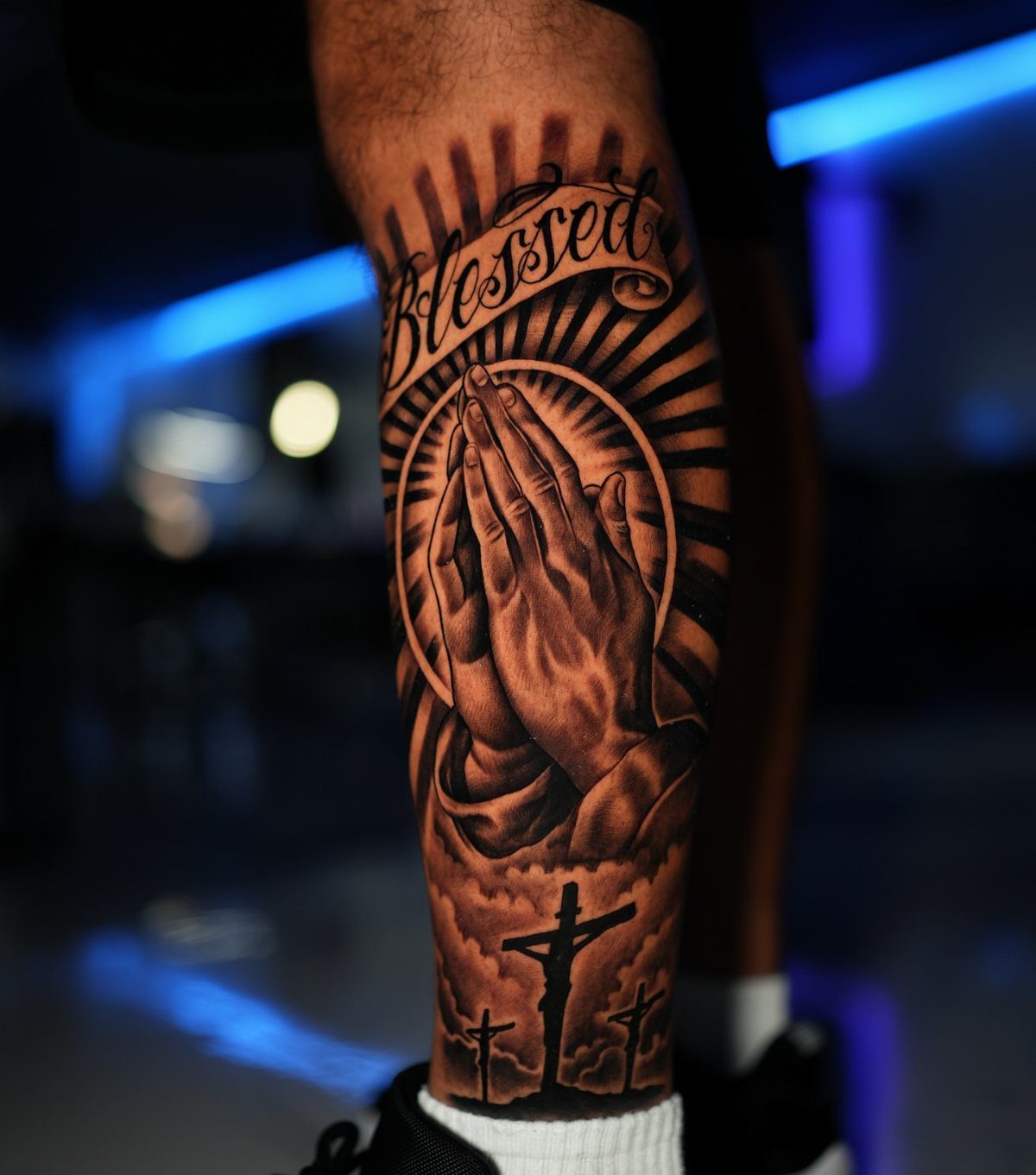 A detailed black and grey tattoo of praying hands and a crucifix on a person's leg.