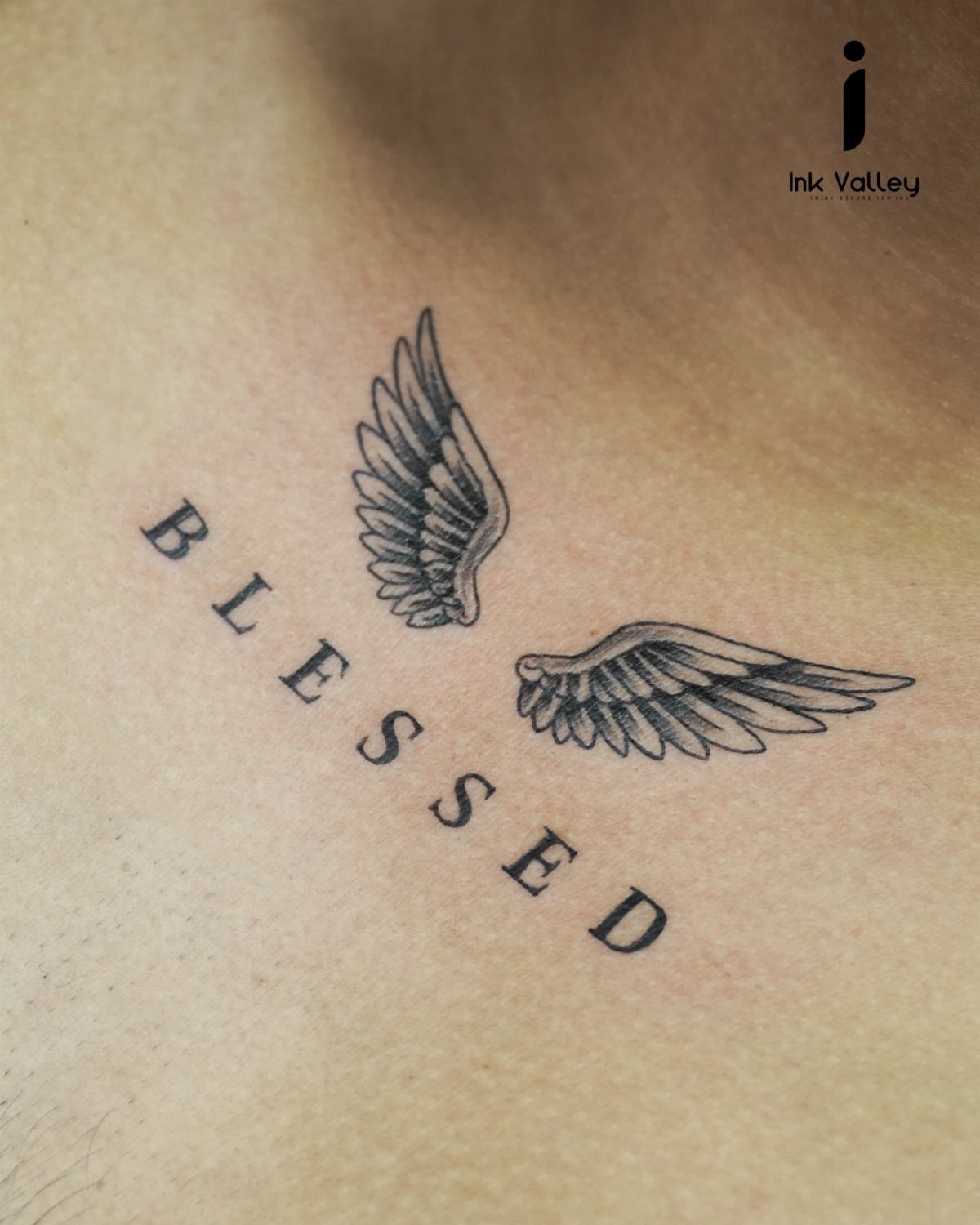 Black ink tattoo of the word BLESSED between two feathered angel wings on skin.