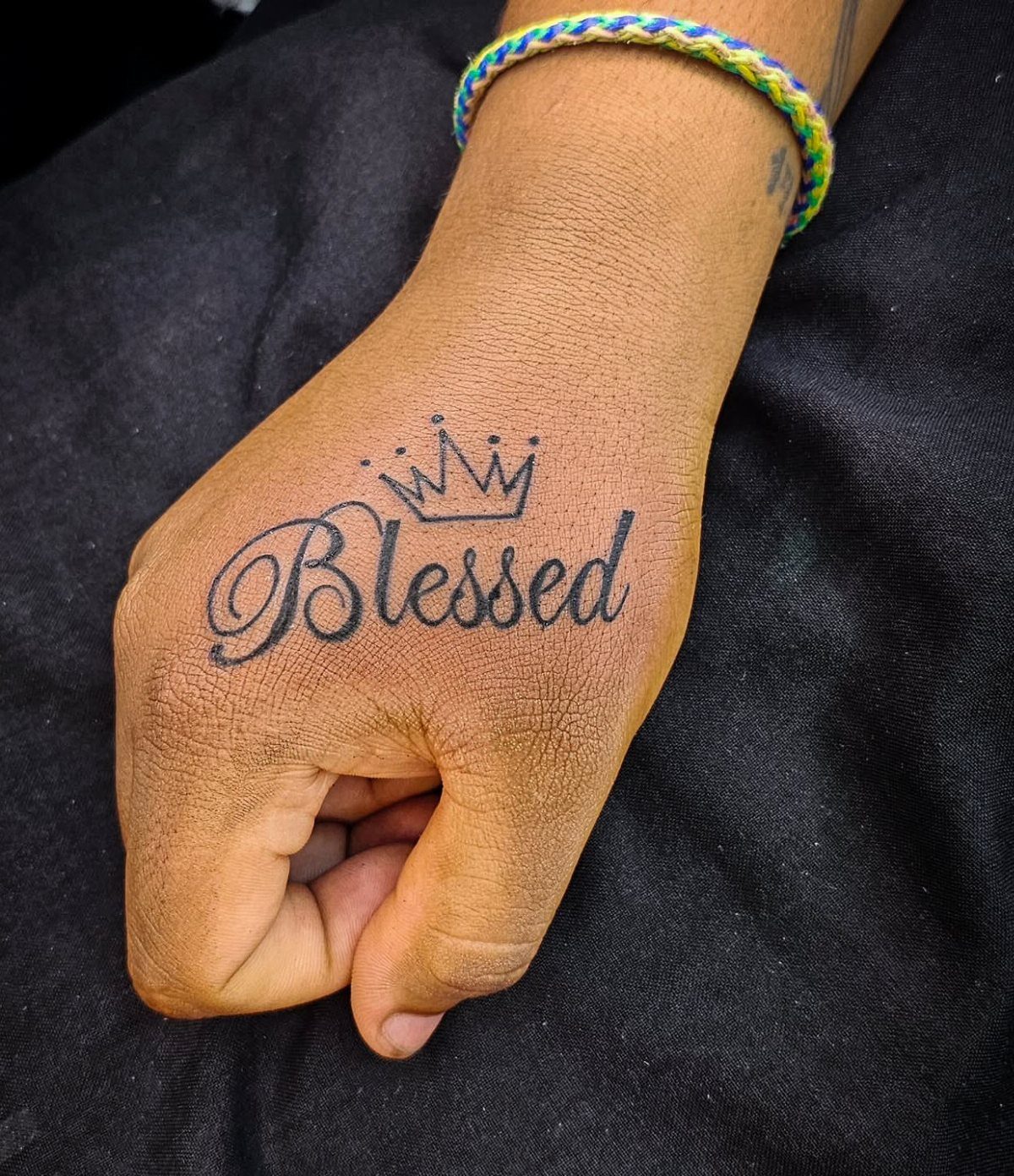 A hand tattoo featuring the word Blessed in cursive script with a small crown above it.