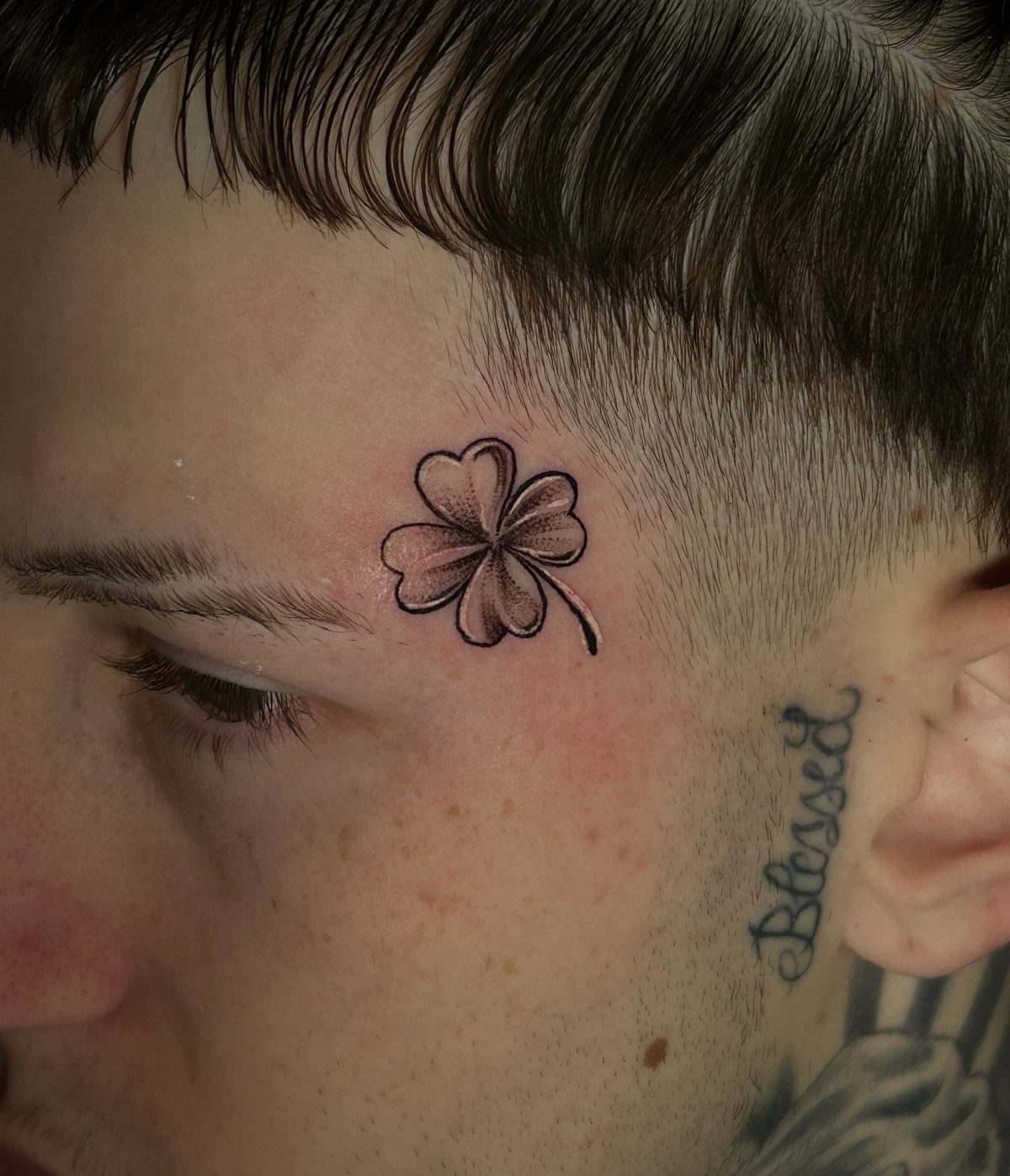 A person's face featuring a small four-leaf clover tattoo and a Blessed script tattoo.