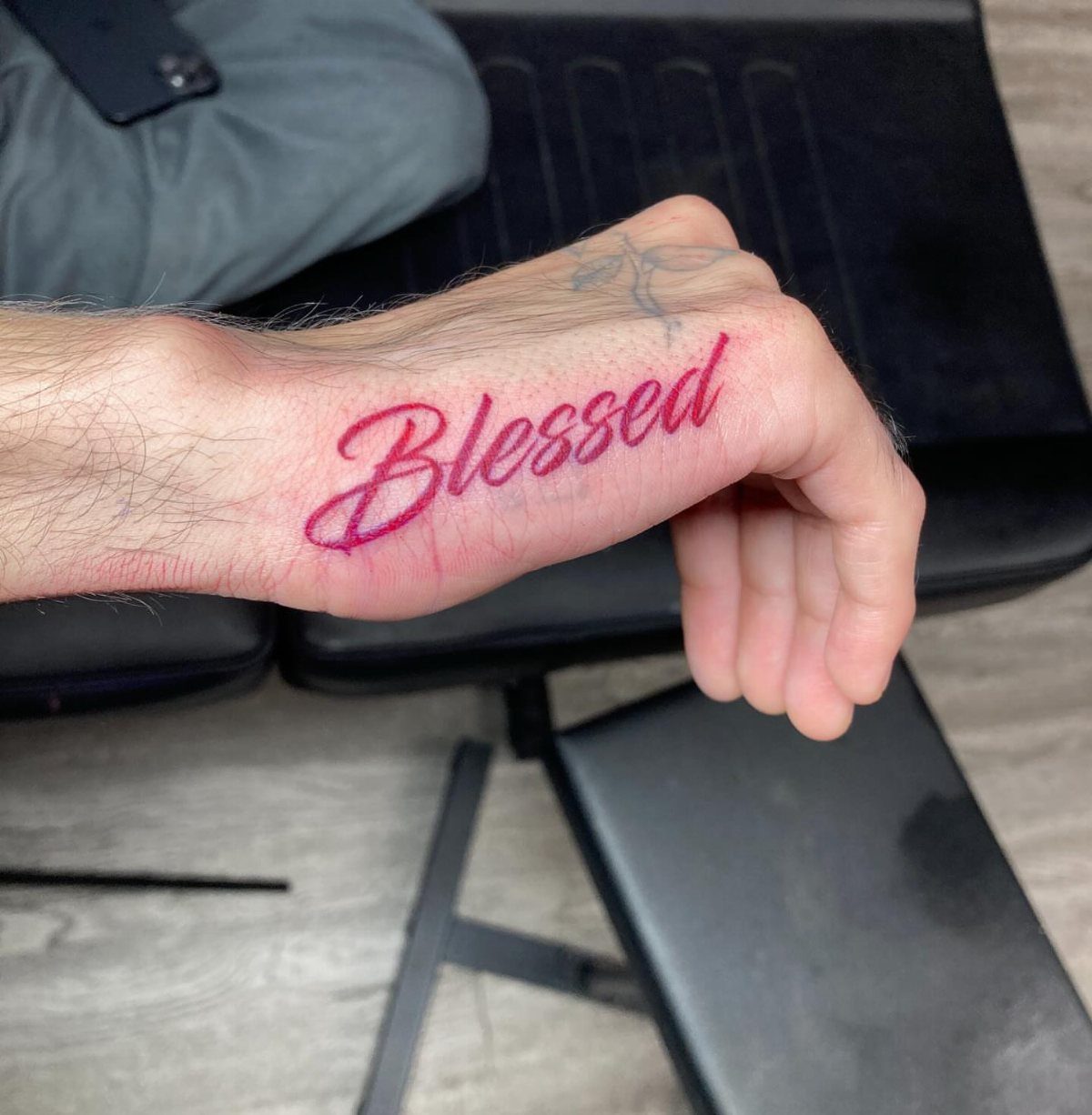 A hand featuring a red ink cursive tattoo of the word Blessed on the side.