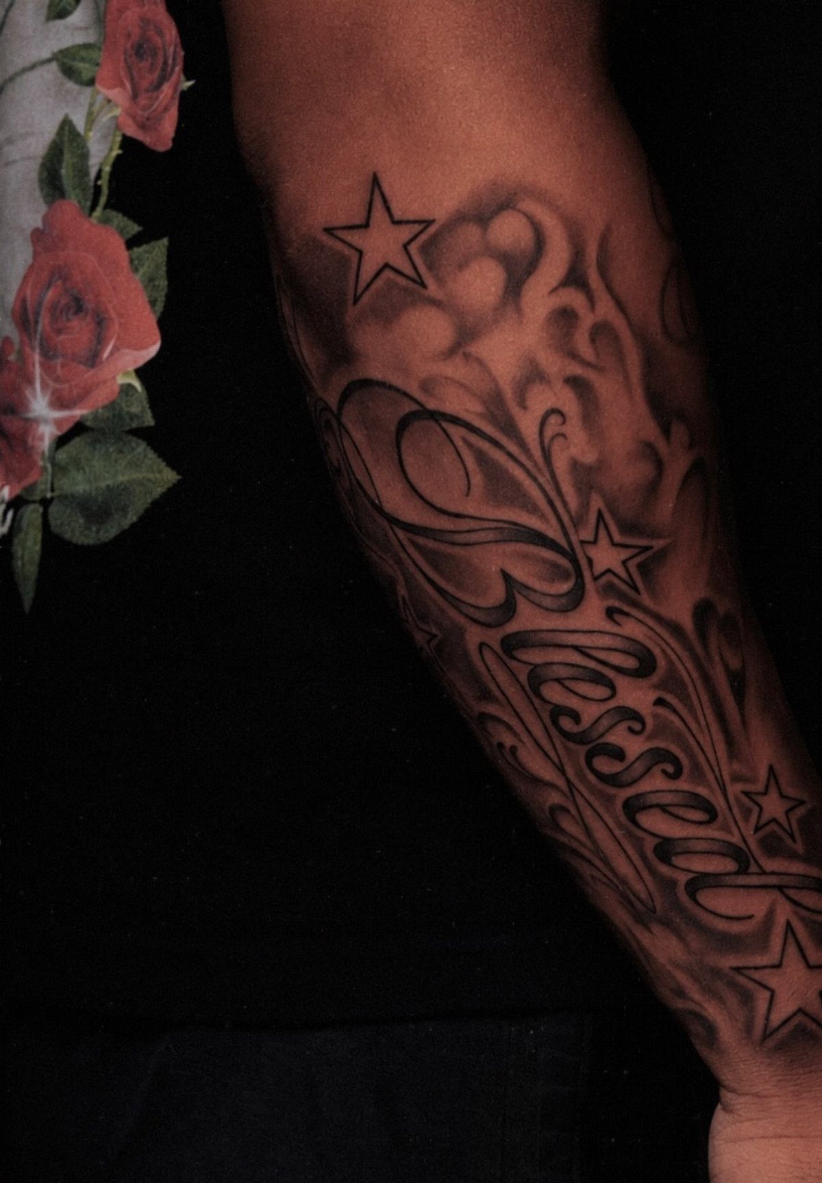 A large forearm tattoo featuring the word Blessed in script with stars and ornamental shading.
