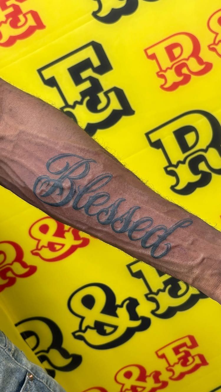 A forearm tattoo featuring the word Blessed in elegant black cursive script lettering.