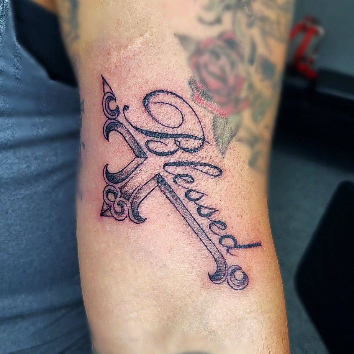 A black ink tattoo on an arm combining the word Blessed with a stylized cross design.