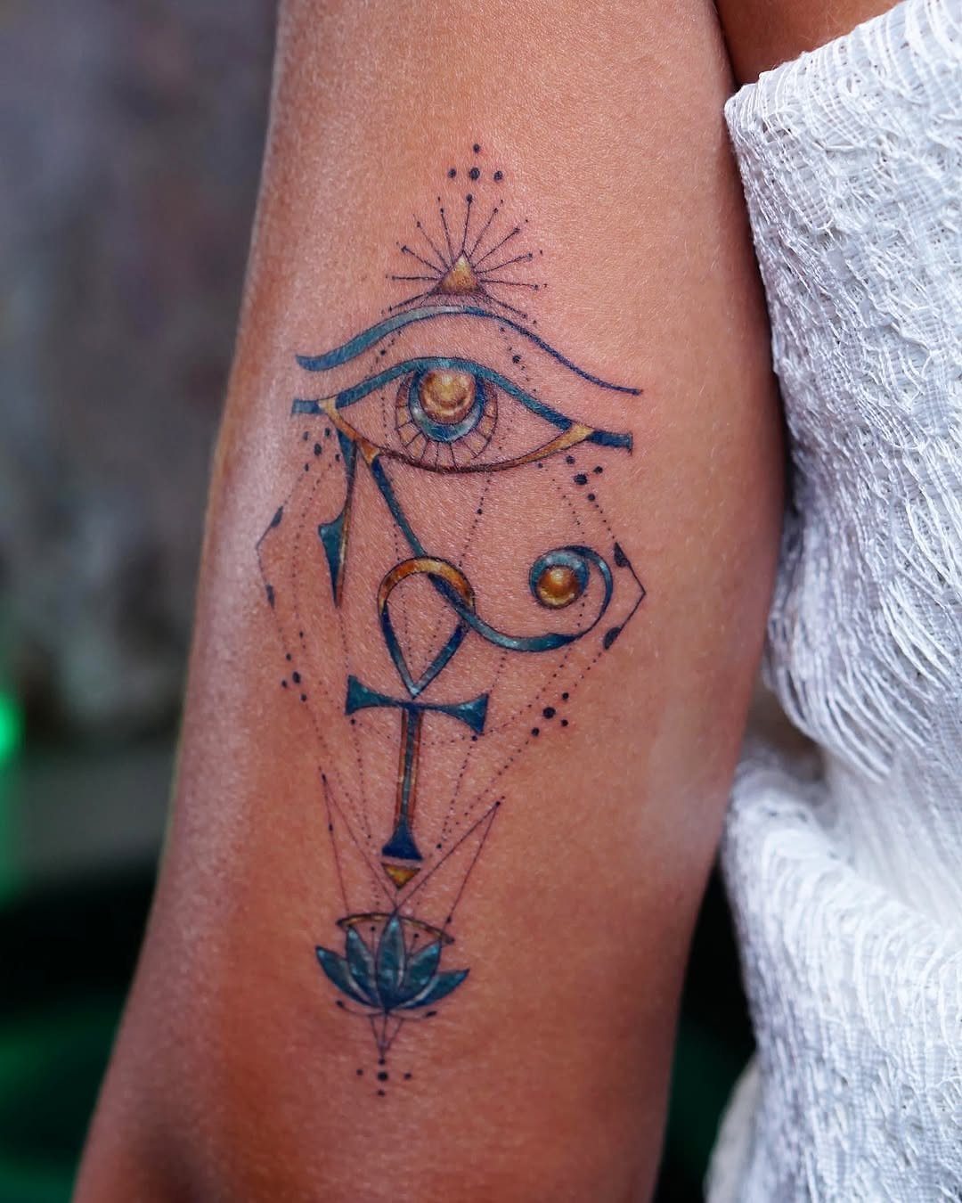 A vertical tattoo on an arm featuring the Eye of Horus, an ankh, and a blue lotus.