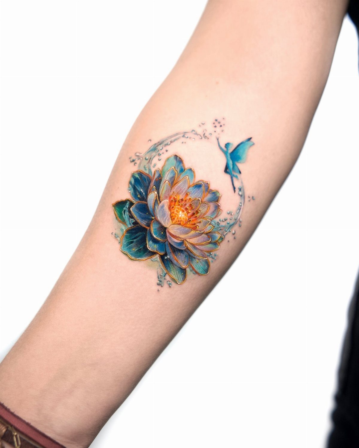 A vibrant blue and orange lotus flower tattoo with a small fairy on a person's forearm.