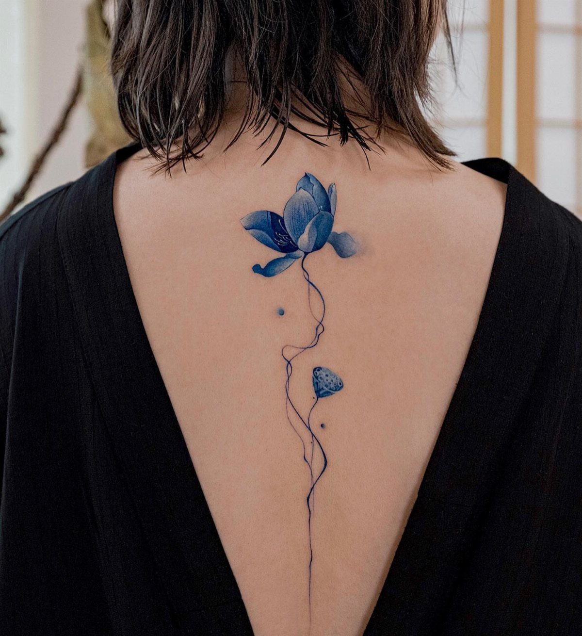 A blue lotus tattoo with a long stem on a person's upper back.