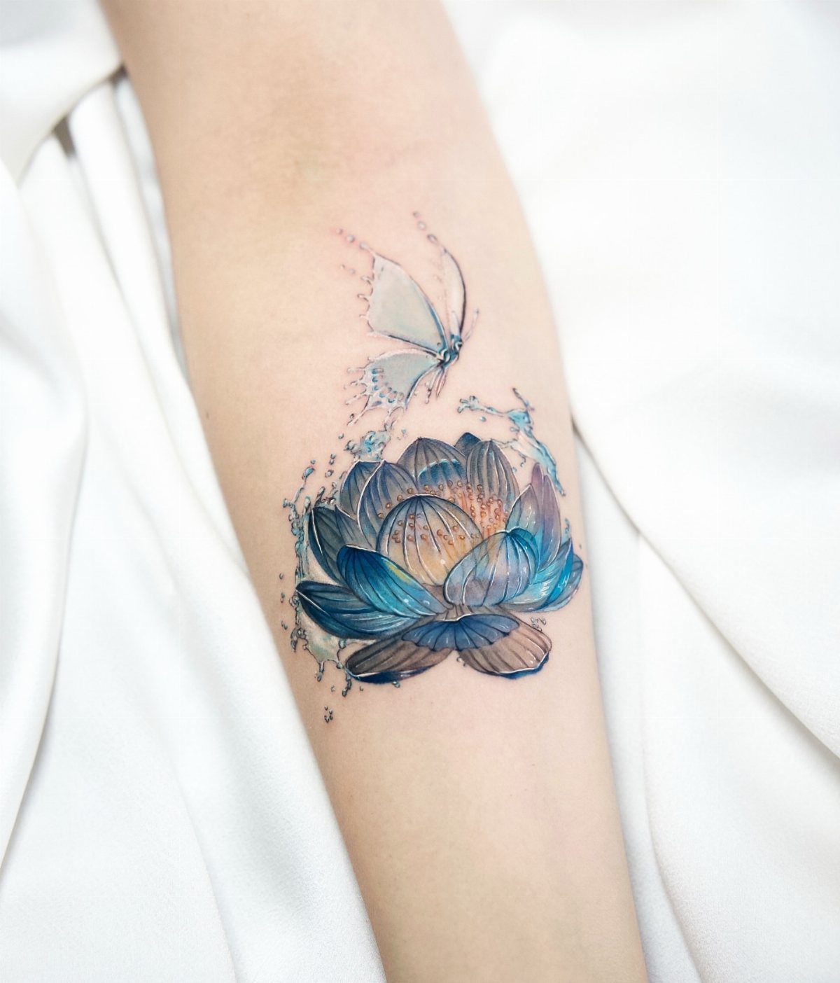 A blue lotus flower and butterfly watercolor style tattoo on a person's arm.