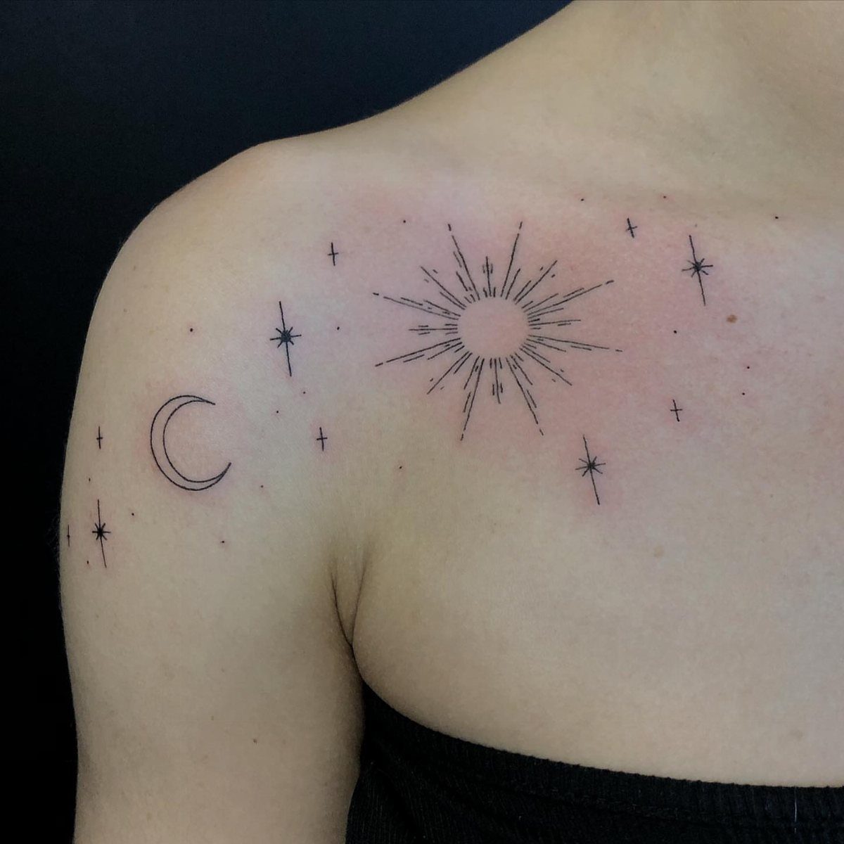 Fine line black ink tattoos of a moon, sun, and stars on a person's collarbone.