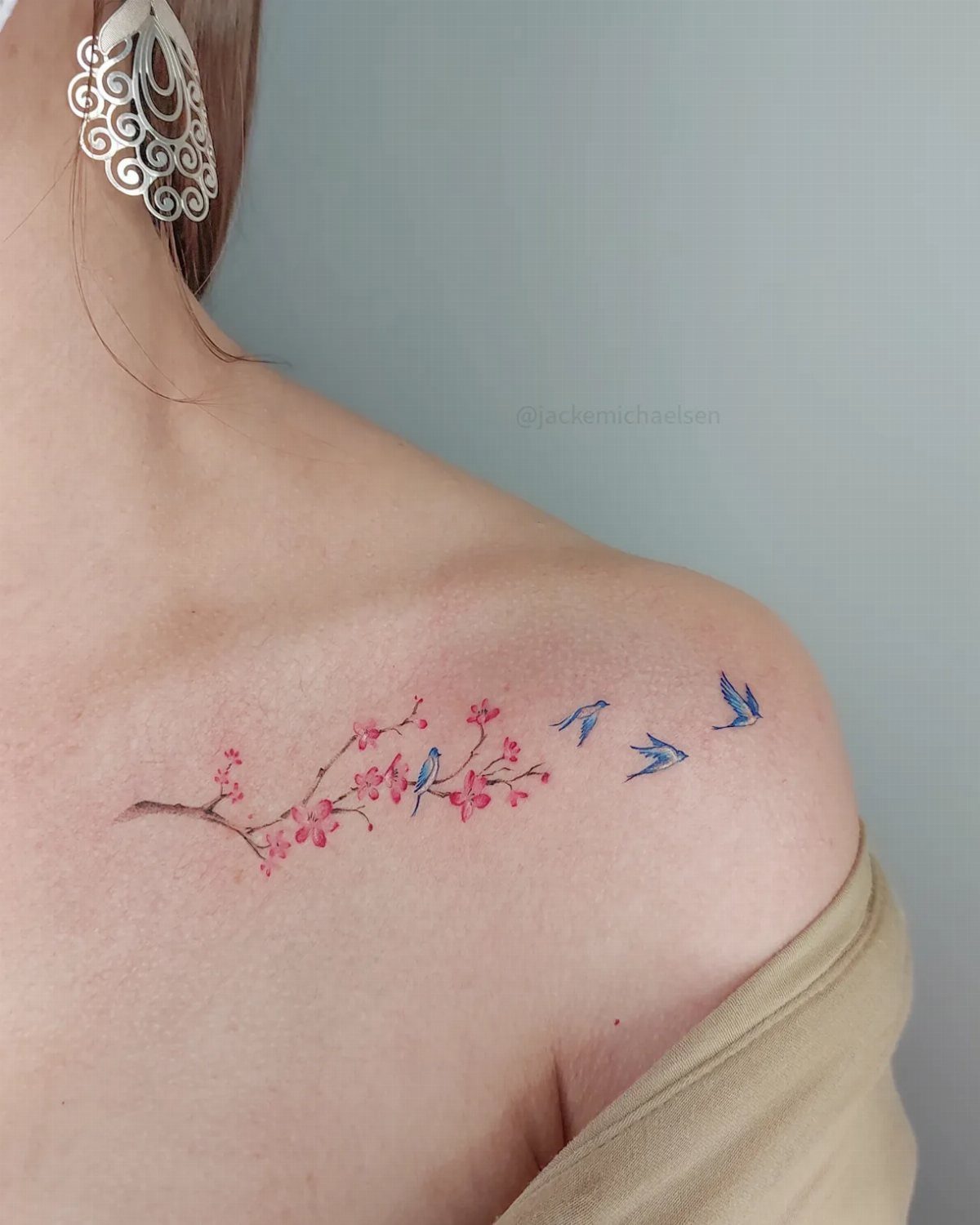 A collarbone tattoo featuring a pink cherry blossom branch and small blue birds in flight.