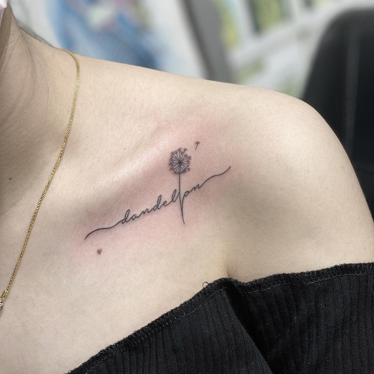 A fine-line black ink dandelion tattoo with cursive text on a person's collarbone.