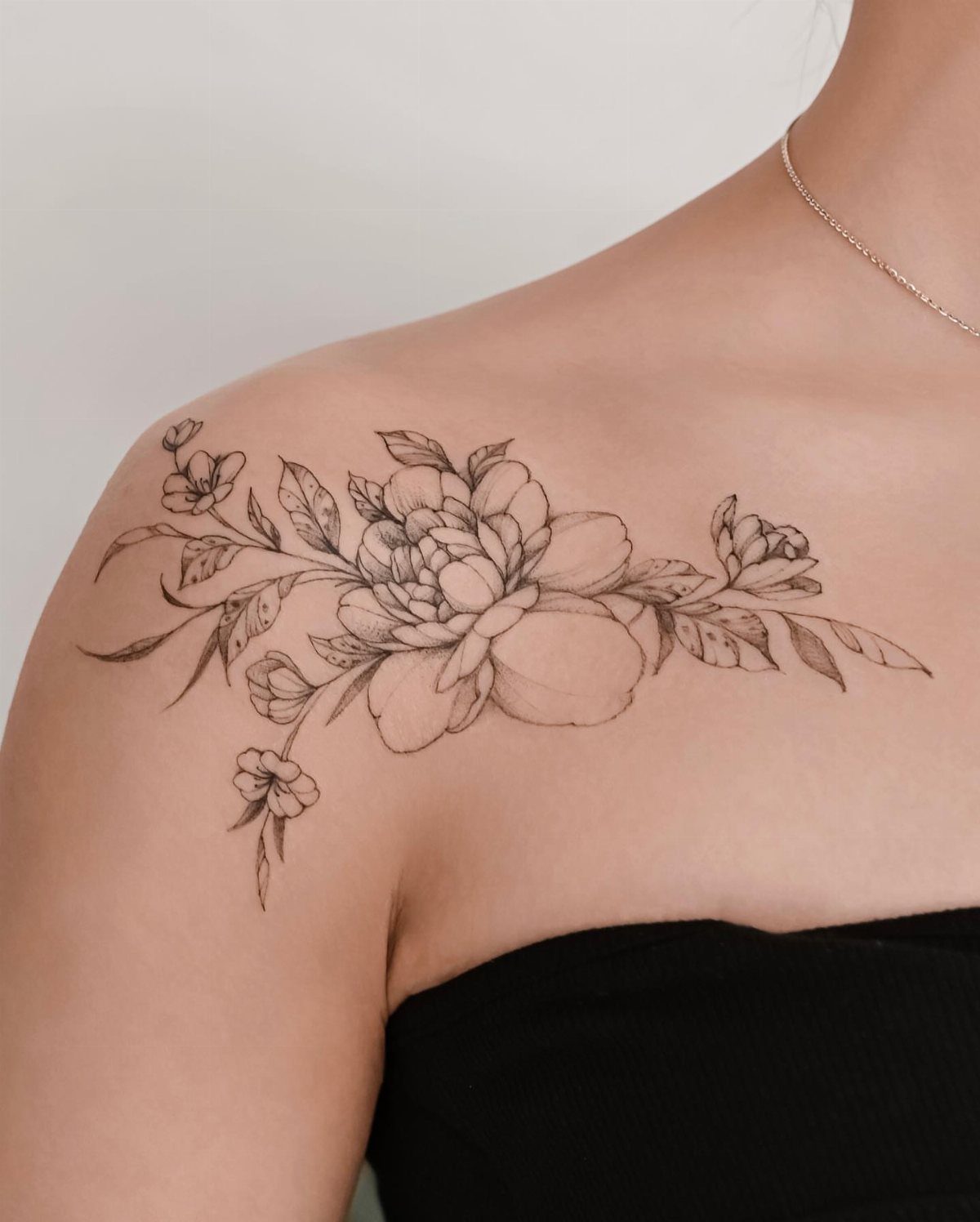 A black ink floral tattoo with peonies and leaves stretching across a person's collarbone.