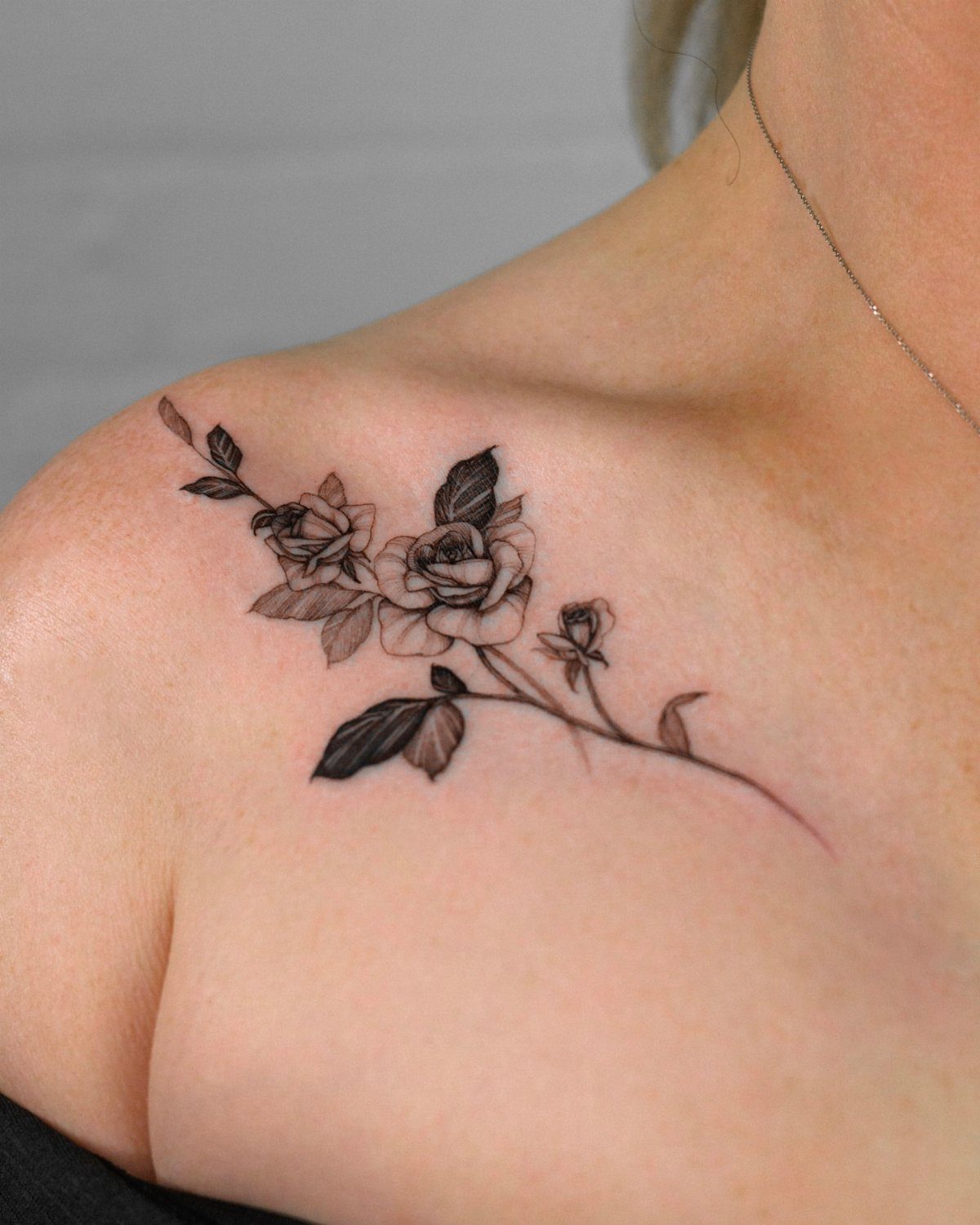 A black and grey rose tattoo with leaves on a person's collarbone and shoulder.