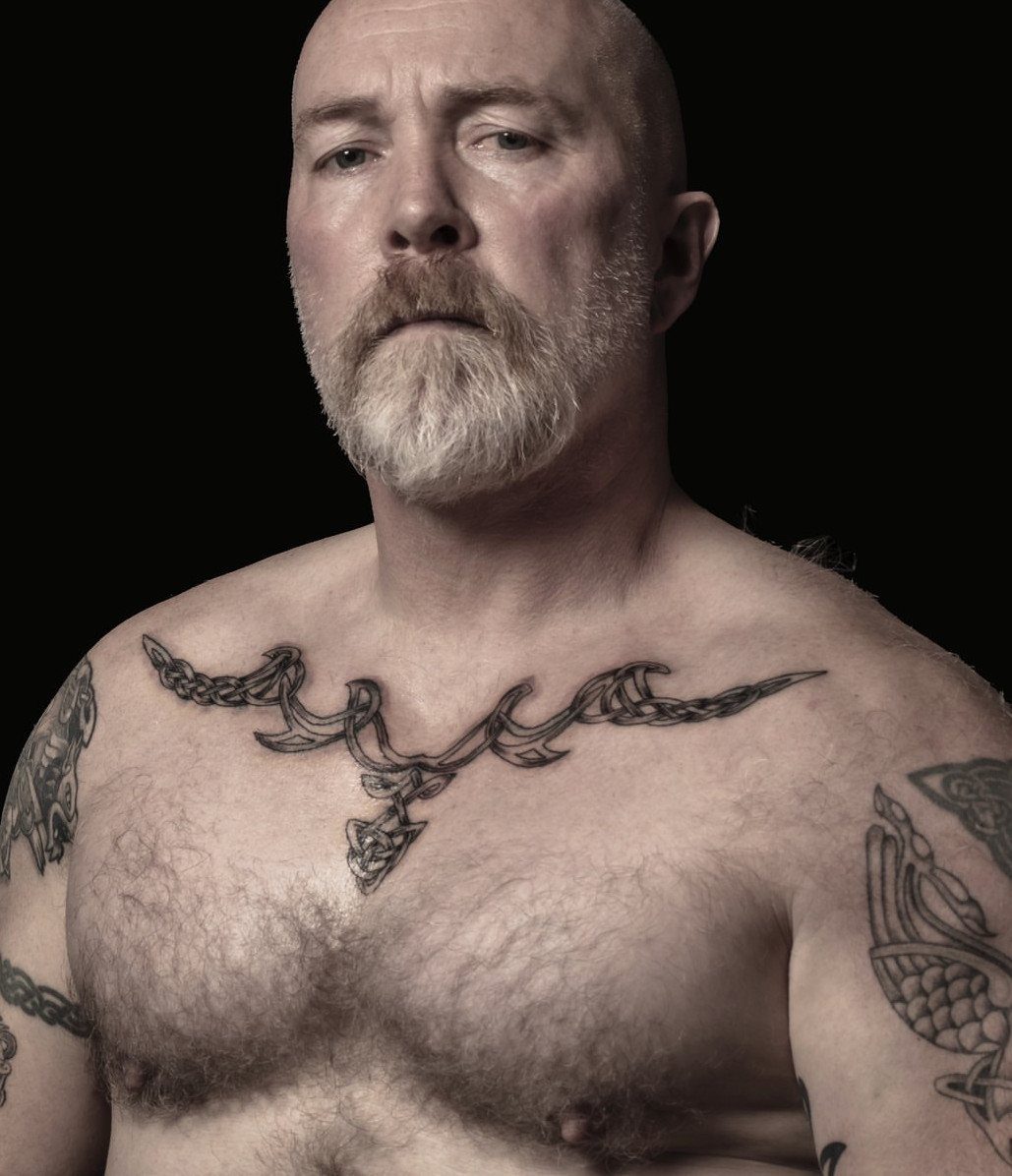 A bald man with a white beard displaying decorative black ink tattoos across his chest and collarbone.