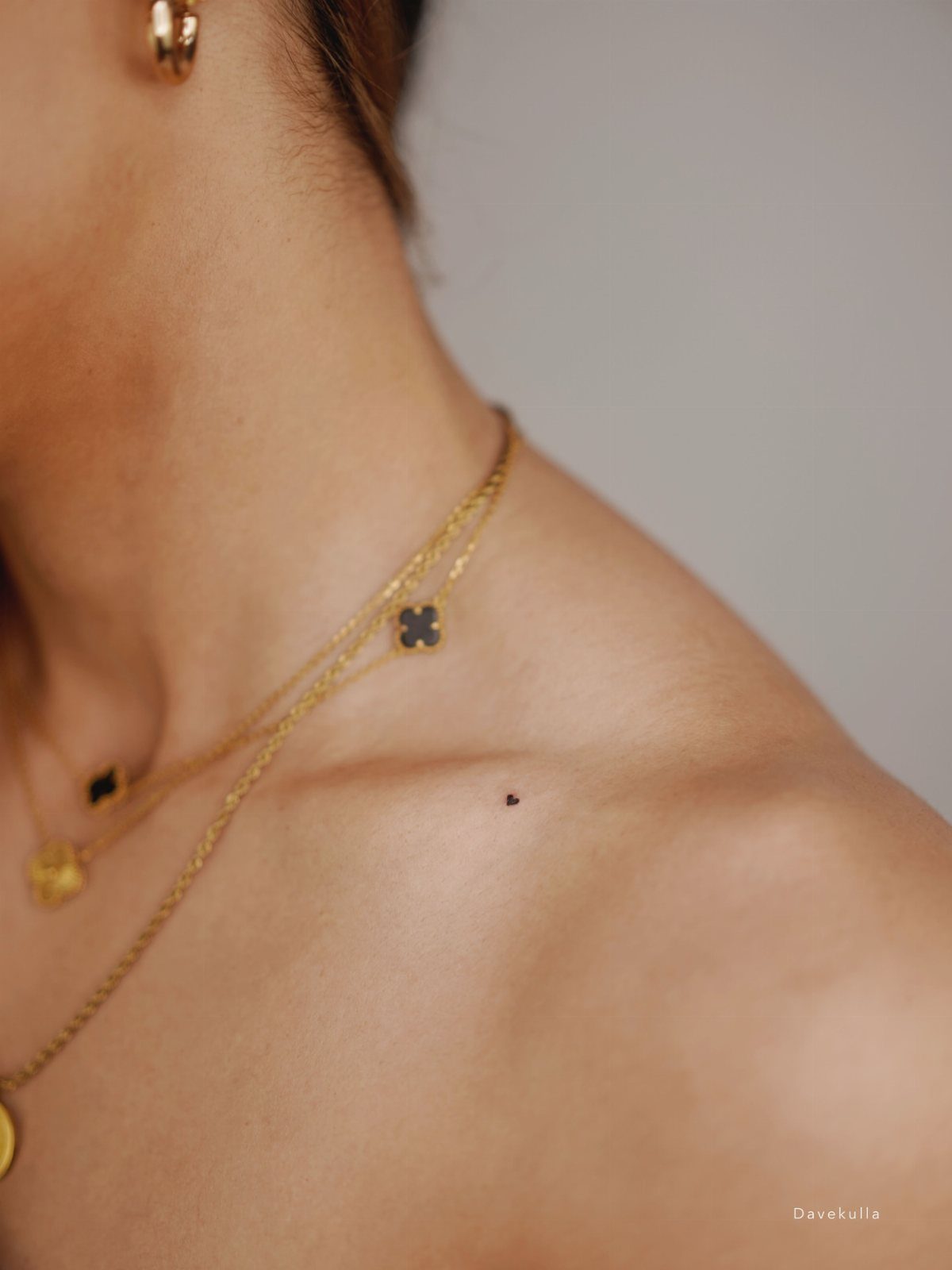 A tiny black heart tattoo located on a person's collarbone near gold necklaces.