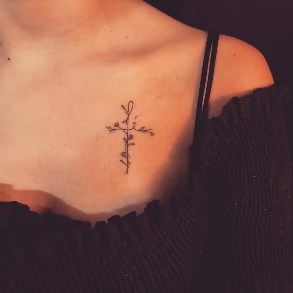 Small floral cross tattoo on a person's collarbone area wearing a black top