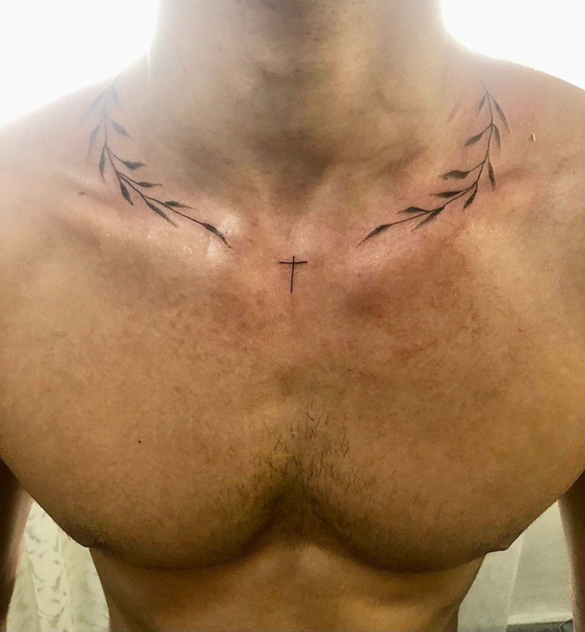 Small black cross tattoo on chest flanked by two leafy branch tattoos on collarbones