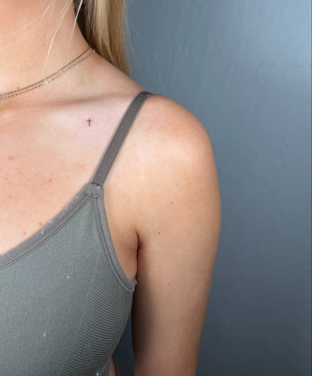 Small black cross tattoo on a person's collarbone wearing a grey tank top.