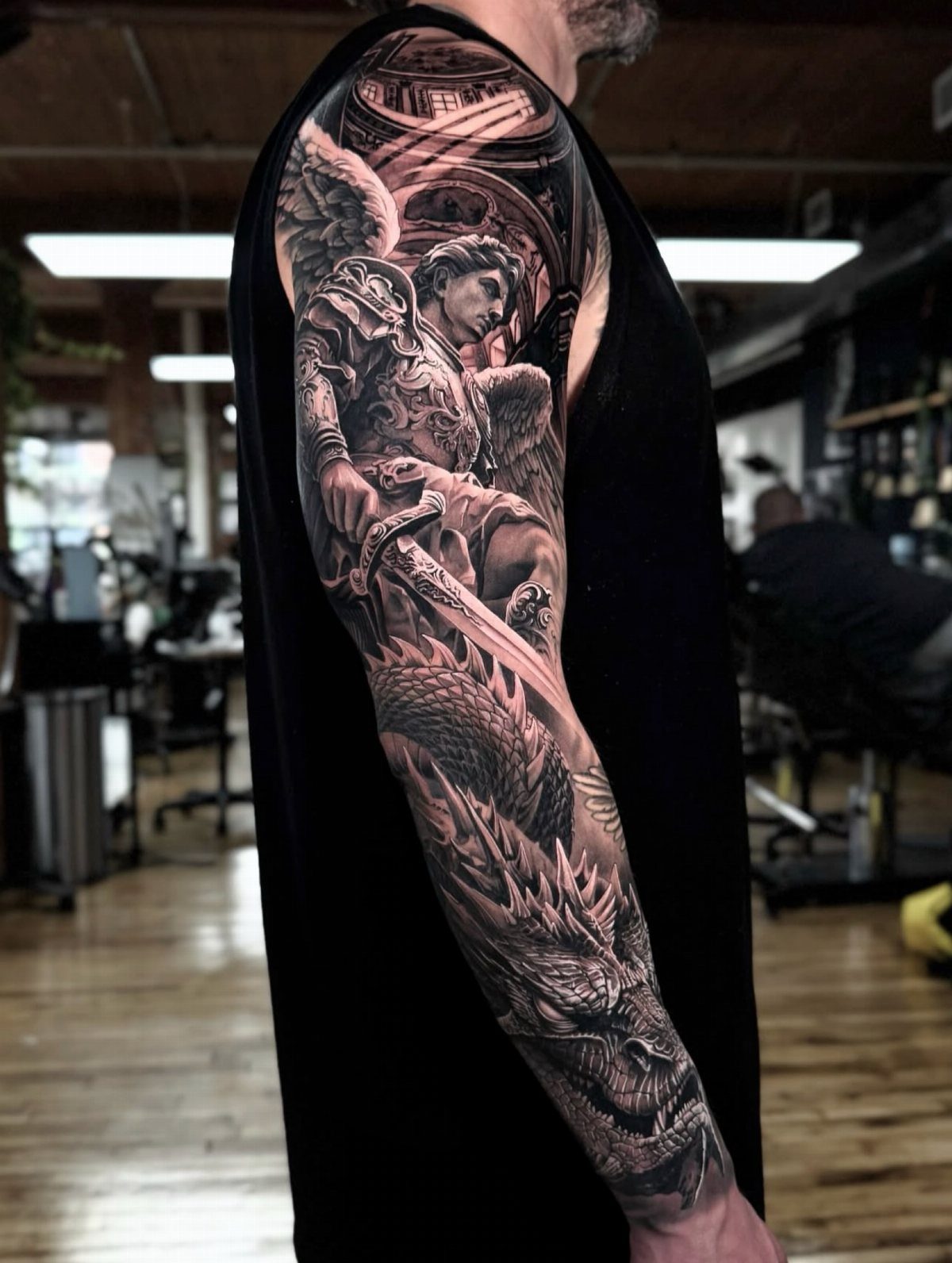 6 Evil Angel Tattoo Design Ideas and Inspiration 6 A detailed black and grey tattoo of a muscular winged angel and a dragon on a man's arm.