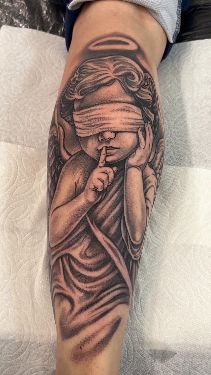 6 Evil Angel Tattoo Design Ideas and Inspiration 2 Black and grey tattoo of a blindfolded cherub angel making a silence gesture on a leg.