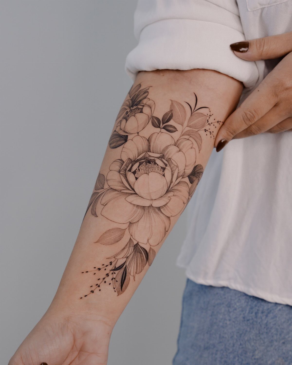 Feminine Fine Line Tattoo: 7 Ideas for Your Next Piece 1 A large black ink floral tattoo of a peony with leaves on a person's forearm.