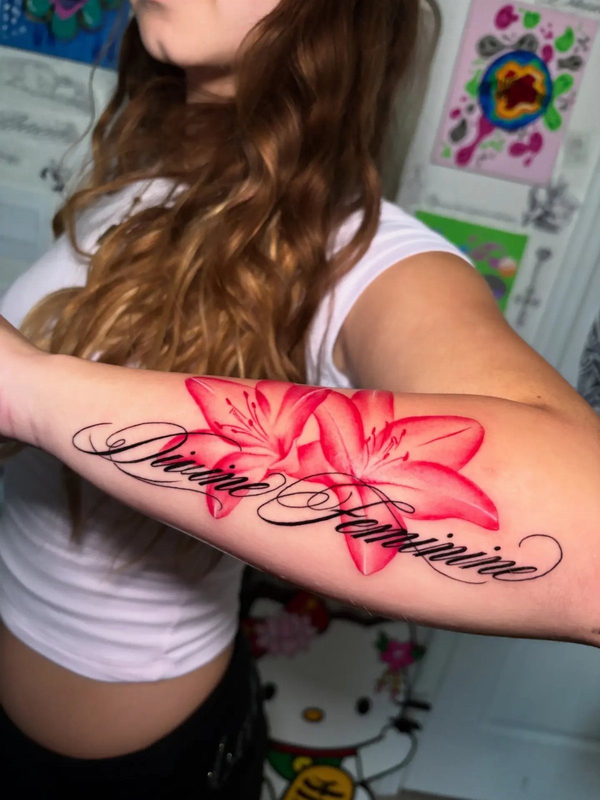 Feminine Fine Line Tattoo: 7 Ideas for Your Next Piece 5 A forearm tattoo featuring two bright pink lilies and the cursive words Divine Feminine.
