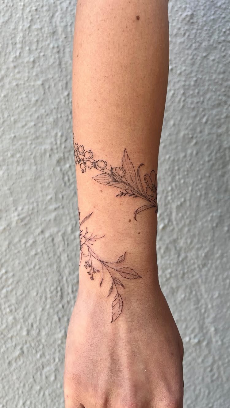 Black ink botanical vine tattoo with delicate leaves and small flowers wrapping around a person's forearm.