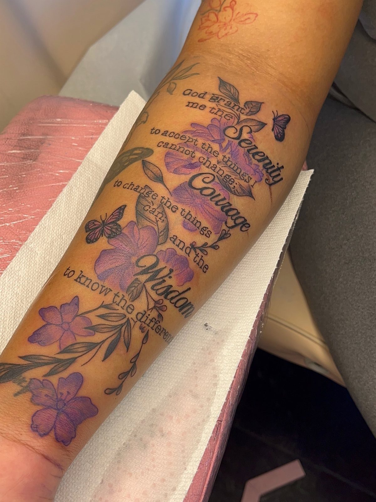A forearm tattoo with purple flowers, butterflies, and cursive script text on skin.