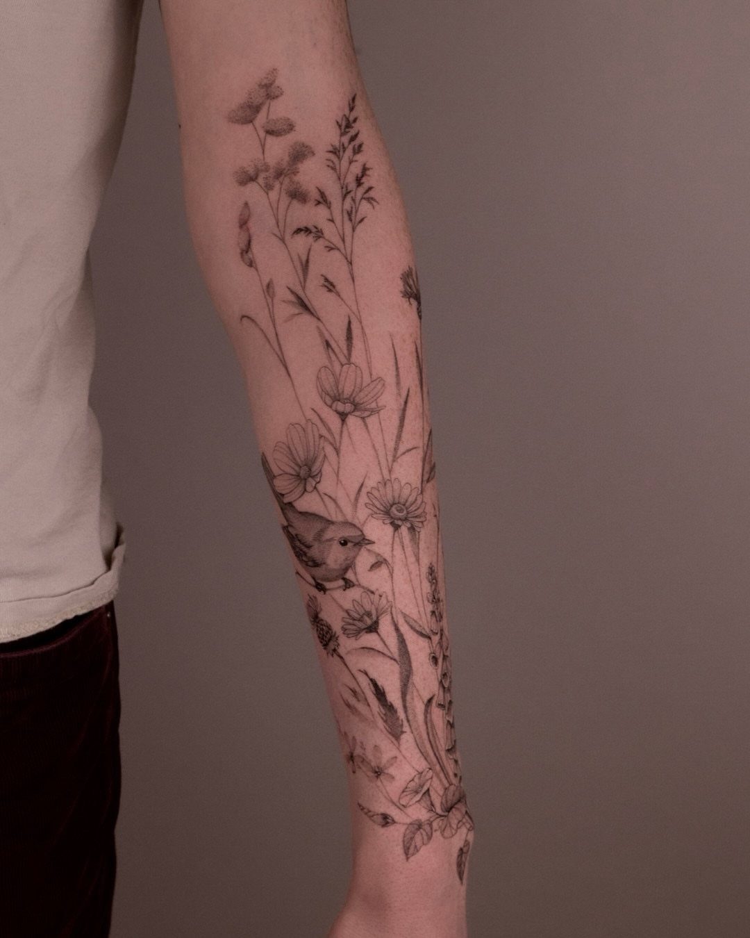 A forearm with a detailed black ink tattoo of delicate wildflowers and a small bird.