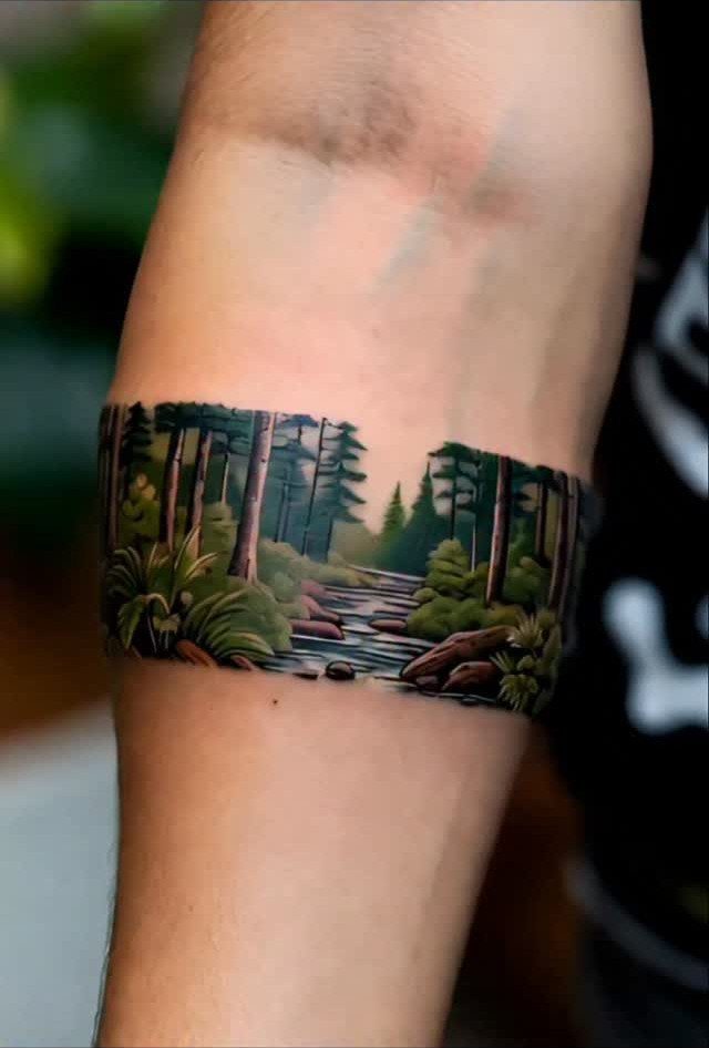 11 Forest Theme Tattoo Ideas: Wolf, Mountain, Sleeve & More 4 A detailed forest landscape tattoo wrapping around a person's forearm with trees and a stream.