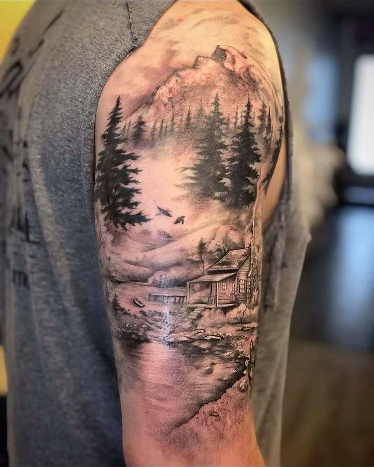 11 Forest Theme Tattoo Ideas: Wolf, Mountain, Sleeve & More 3 A detailed black and grey mountain and forest landscape tattoo on a person's upper arm.