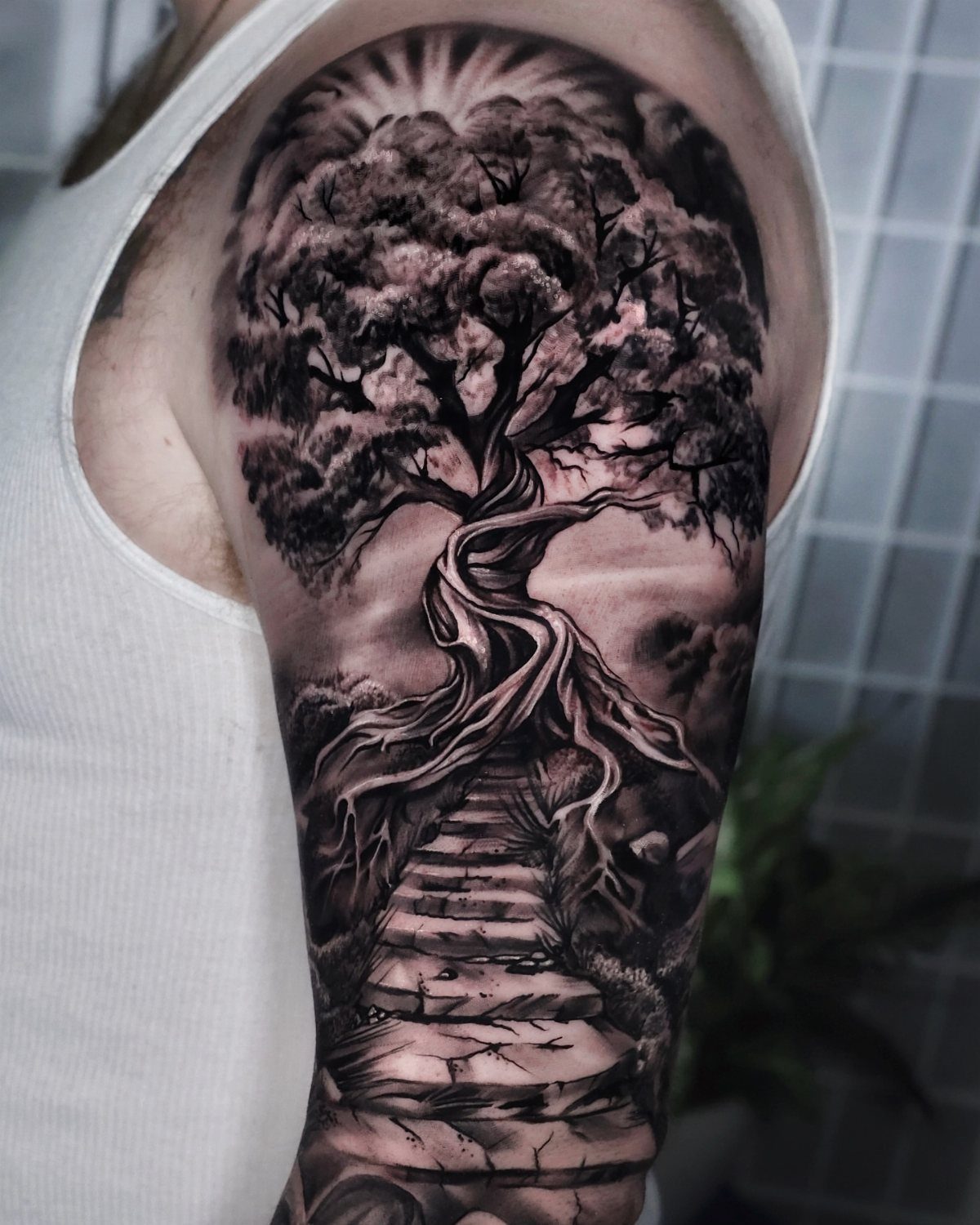 11 Forest Theme Tattoo Ideas: Wolf, Mountain, Sleeve & More 10 A detailed black and grey tattoo of a large gnarled tree with stone steps on an arm.