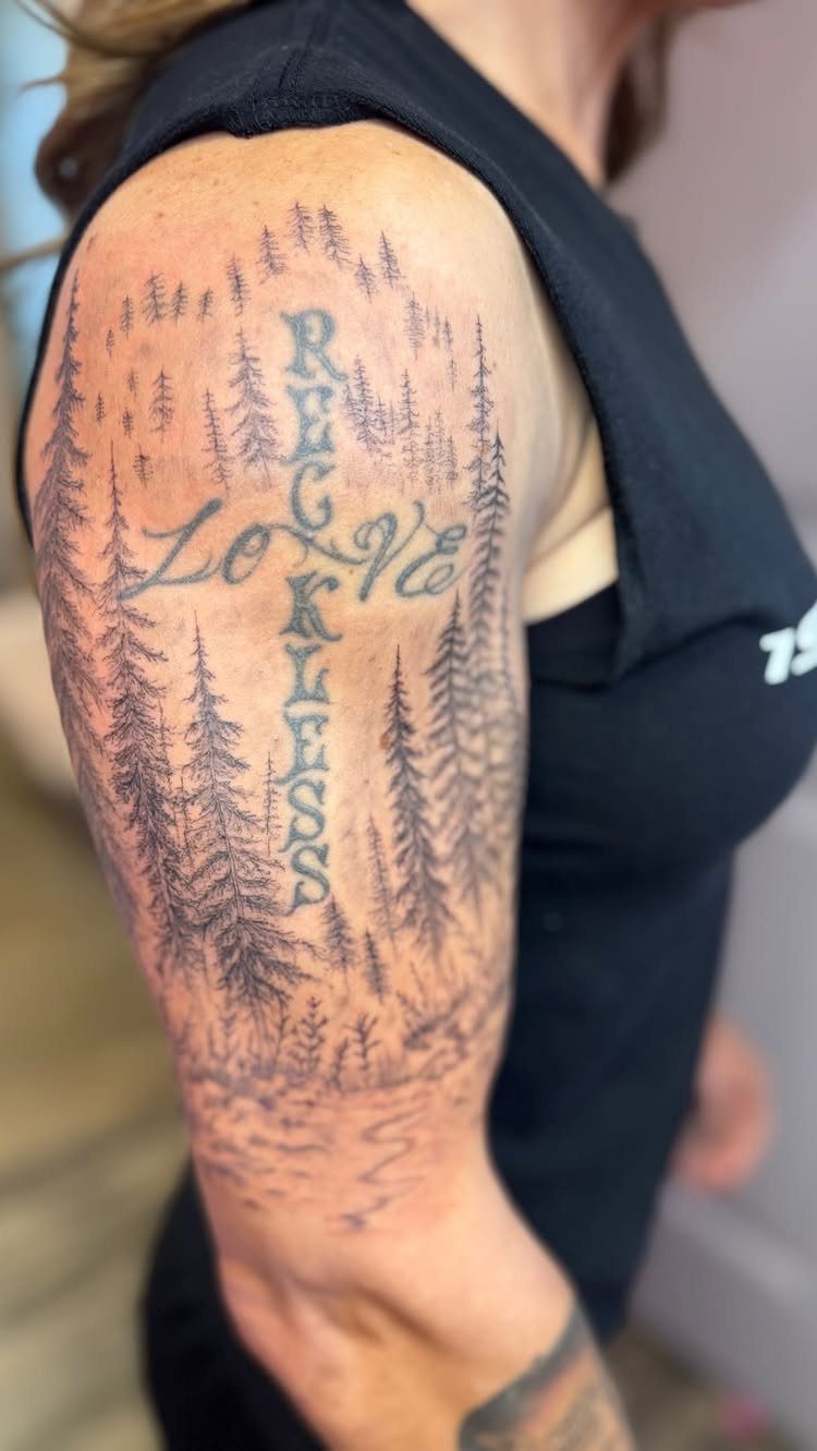 11 Forest Theme Tattoo Ideas: Wolf, Mountain, Sleeve & More 11 A large black ink forest tattoo with pine trees and vertical text on a person's arm.