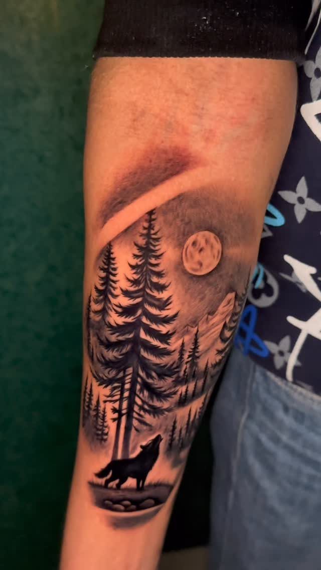 11 Forest Theme Tattoo Ideas: Wolf, Mountain, Sleeve & More 1 A black and grey forearm tattoo depicting a howling wolf silhouette amidst pine trees and a moon.