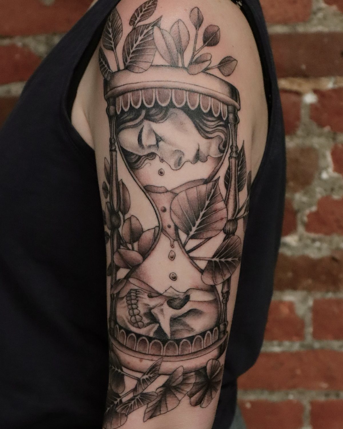 Hour Glass Tattoo Ideas: 12 Designs for Every Style 12 A large black and grey realism tattoo of an hourglass with leaves on a person's arm.
