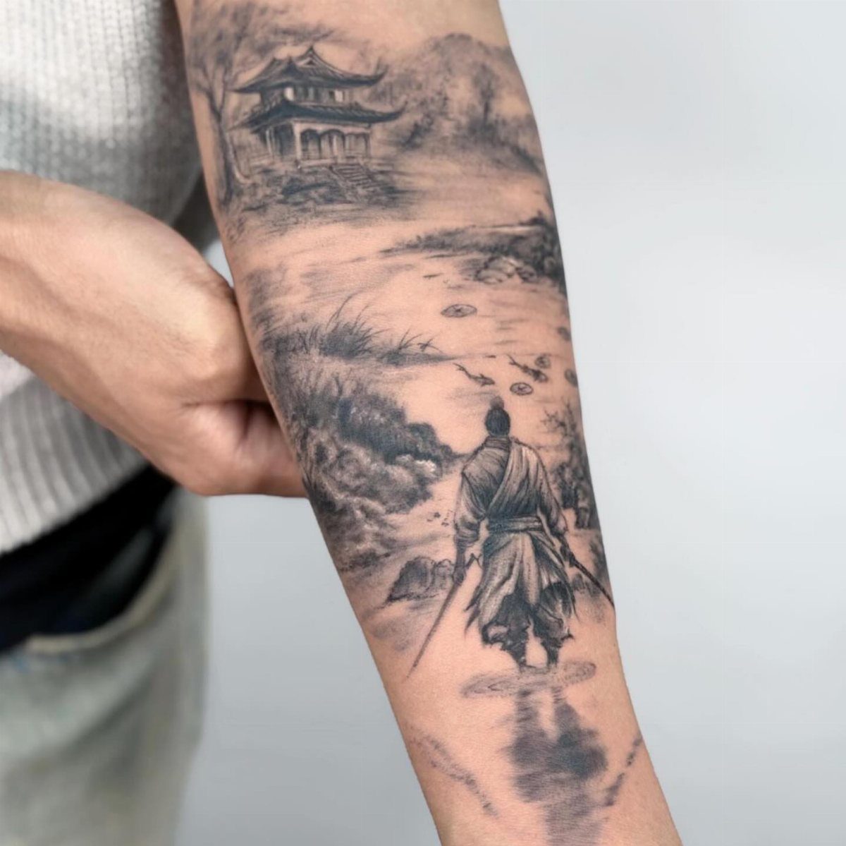 A black and grey landscape tattoo on a forearm featuring a pagoda and mountains.